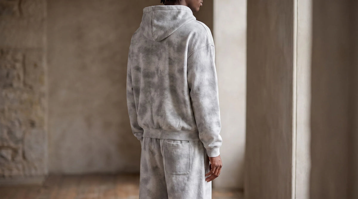Rear three-quarter study of the SM Cloud Fade Hoodie and matching sweatpants beside a softly lit window.