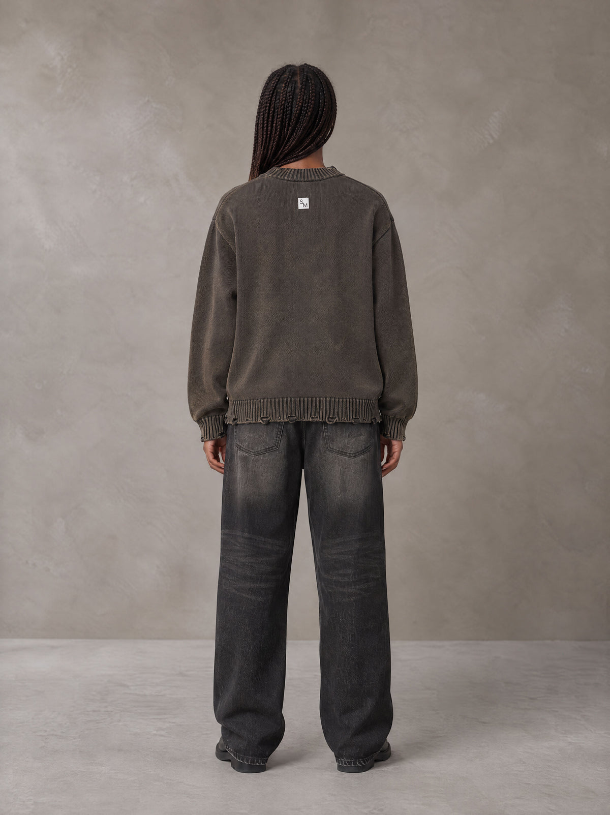 Model wearing the SM Sig. Washed Distress Knit Sweater in Kabul Brown with SM Archive Fade Denim Jeans in black, rear view.