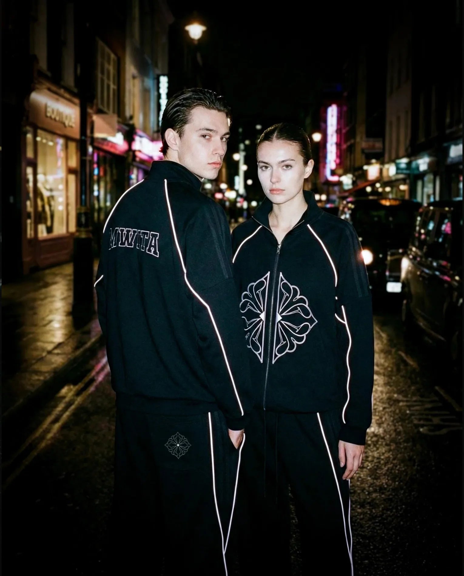Two models wearing SM Fleur Echo Eclipse Two Piece with reflective piping on a city street at night.