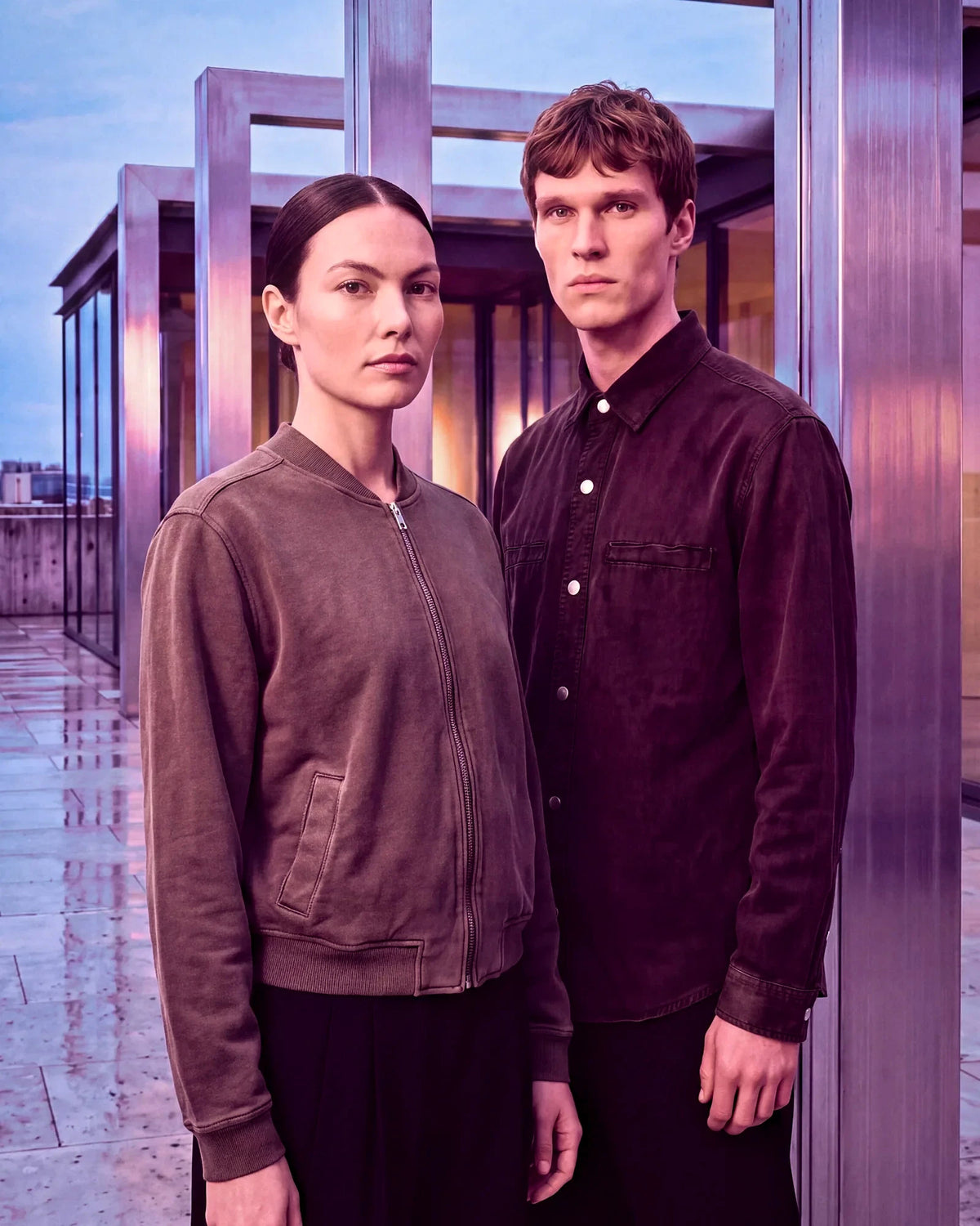 A man and a woman standing on a rooftop terrace at dusk, wearing dark brown and black corduroy and suede jackets. The man wears a dark brown corduroy button-up shirt, and the woman wears a dark brown corduroy zip-up bomber jacket.