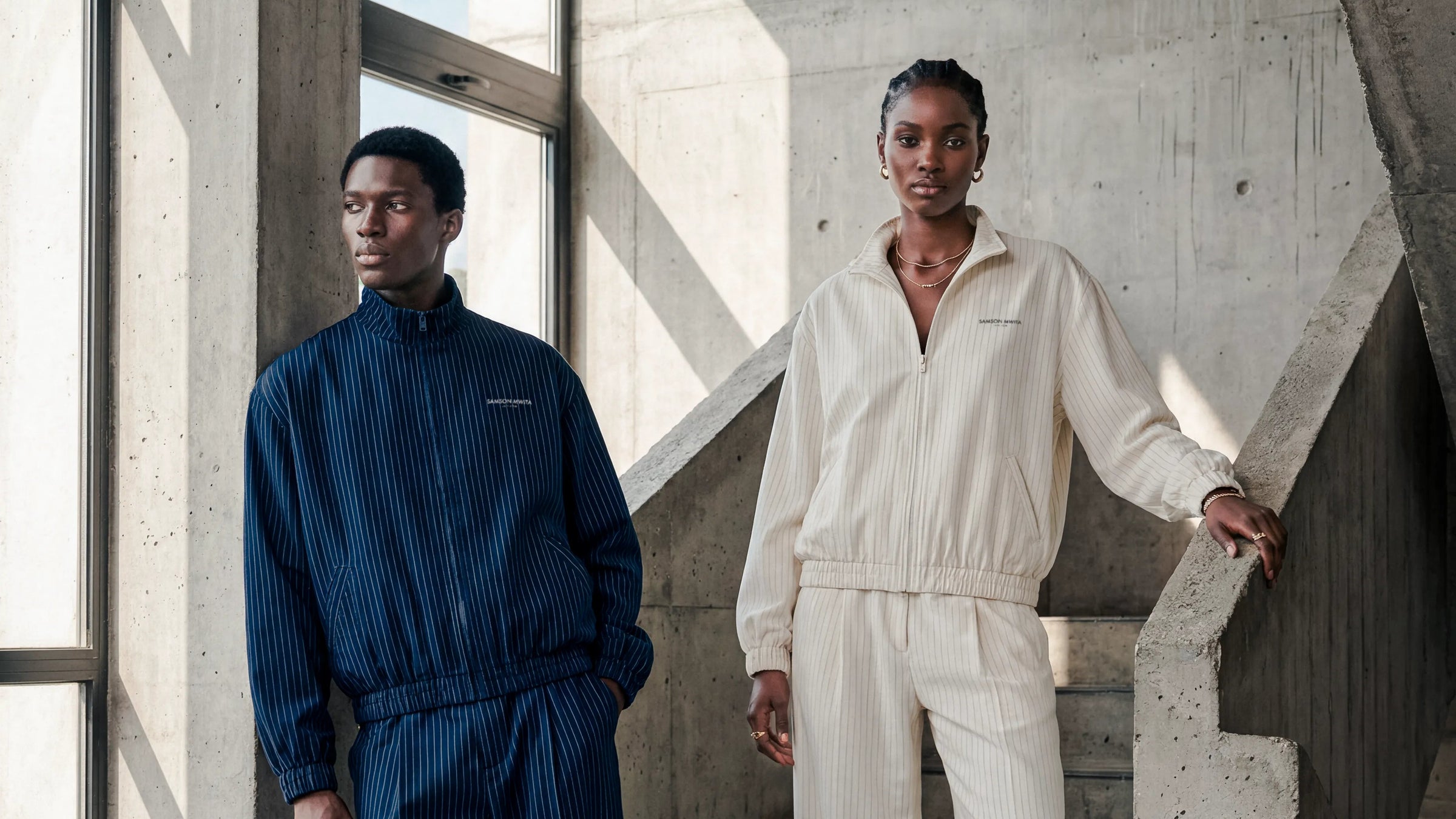 Hero - Two models wearing SM Axis two-piece in a concrete stairwell. One model wears dark navy blue, and the other wears off-white/cream.