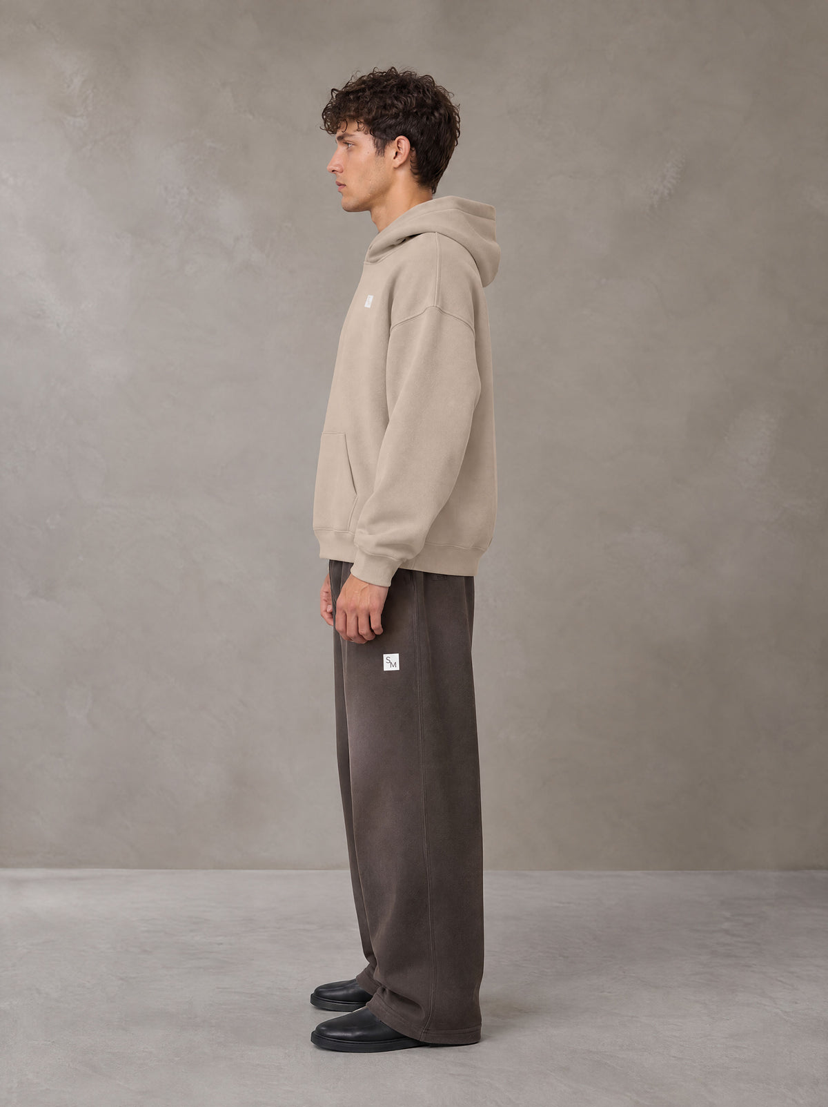 Model wearing SM Sig. Fleece Hoodie in Apricot Grey, side profile.