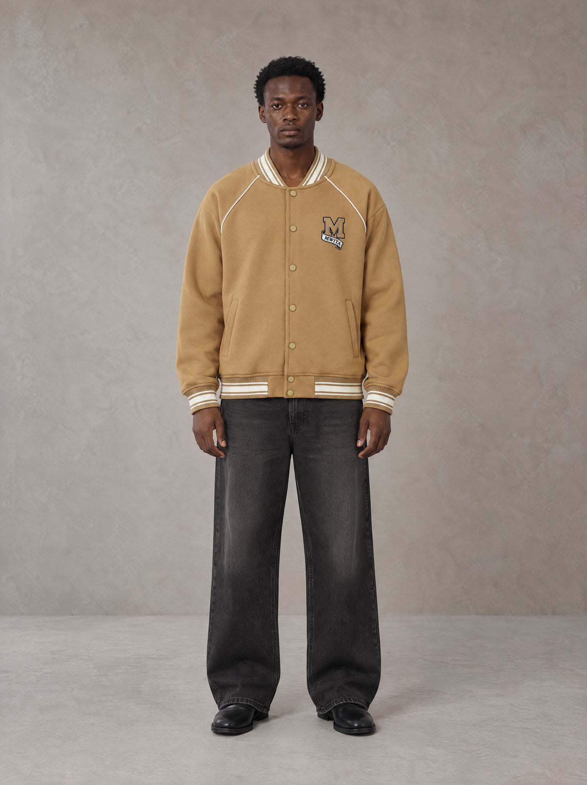 Model wearing the SM Varsity Jacket in Camel Tan, front view.