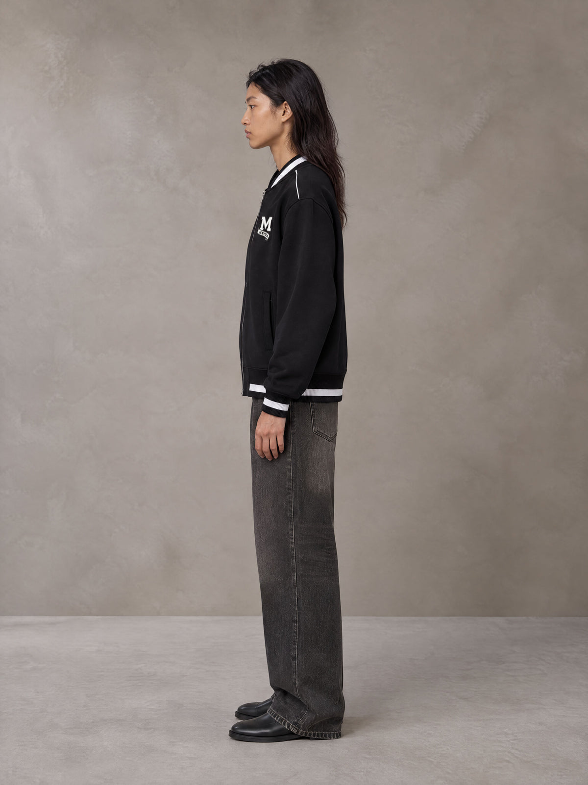 Model wearing the SM Varsity Zip Jacket in Midnight Black, side profile.
