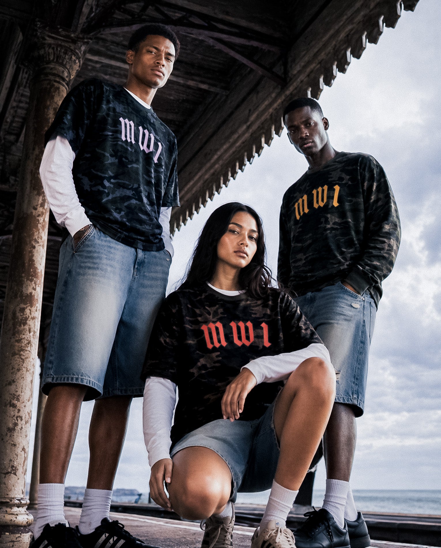 Models wearing matching SM Script Camo T-shirts and Heritage Denim Shorts by the seaside.